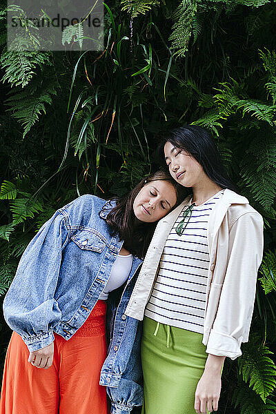 Young lesbian couple with eyes closed leaning on each other in front of plants