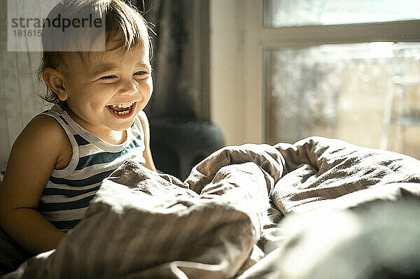 Cute baby laughing in duvet on bed at home