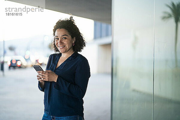 Glückliche Geschäftsfrau mit Mobiltelefon  die an einer Glaswand steht