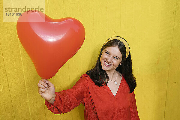 Glückliche junge Frau hält einen herzförmigen Ballon vor einer gelben Wand