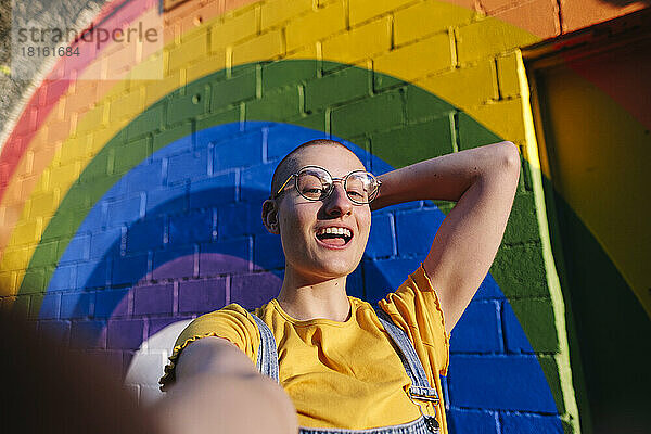 Glückliche androgyne Person mit der Hand hinter dem rasierten Kopf  die ein Selfie vor der Regenbogenwand macht