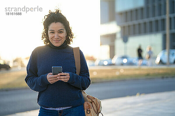 Lächelnde Geschäftsfrau mit Smartphone steht auf Fußweg