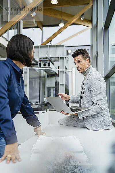 Reifer Geschäftsmann hält Laptop in der Hand und diskutiert mit Kollegen am Schreibtisch in der Industrie