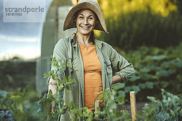 Lächelnde Frau mit der Hand auf der Hüfte und Hut im Garten