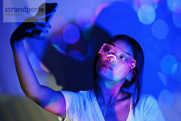 Junge Frau mit futuristischer Brille macht ein Selfie mit dem Handy vor der Wand