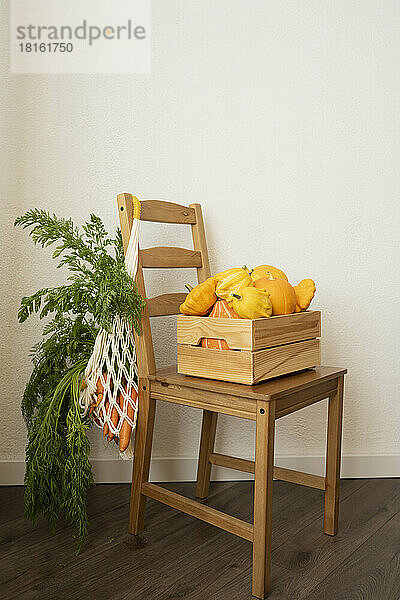 Netzbeutel mit Karotten und Kiste mit Kürbissen auf Stuhl vor Wand