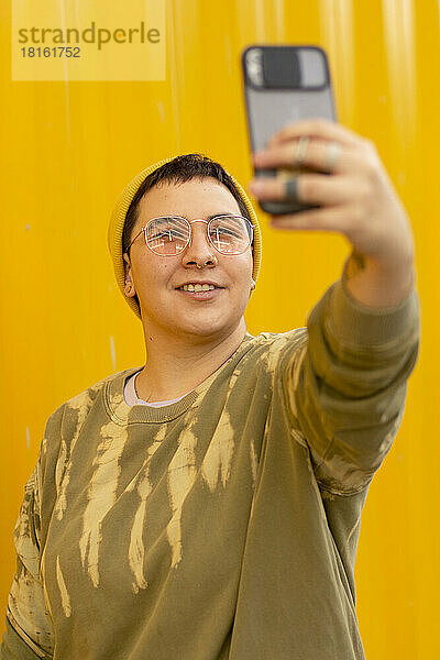 Fröhliche nicht-binäre Person  die vor einer gelben Wellwand ein Selfie mit dem Mobiltelefon macht