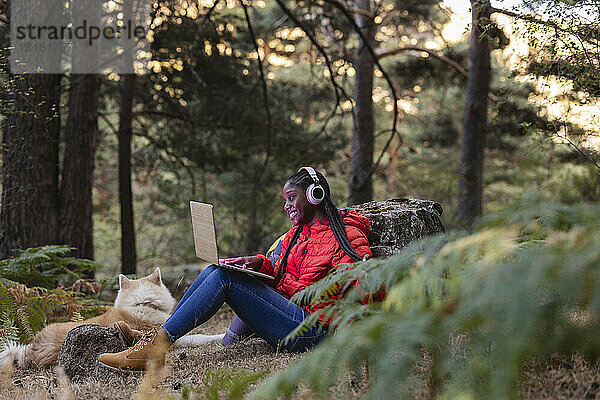 Glückliche Frau mit kabellosen Kopfhörern und Laptop im Wald