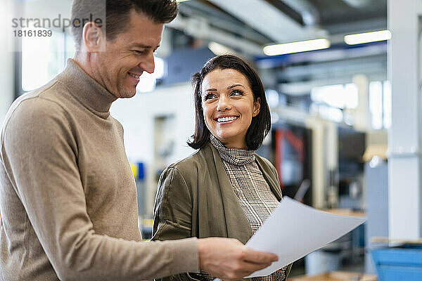 Lächelnde Geschäftsfrau blickt auf Geschäftsmann  der Papier in der Produktionshalle hält