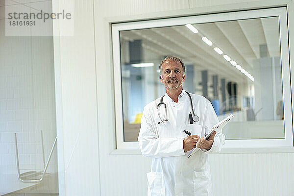 Leitender Arzt mit Klemmbrett steht vor Glasfenster