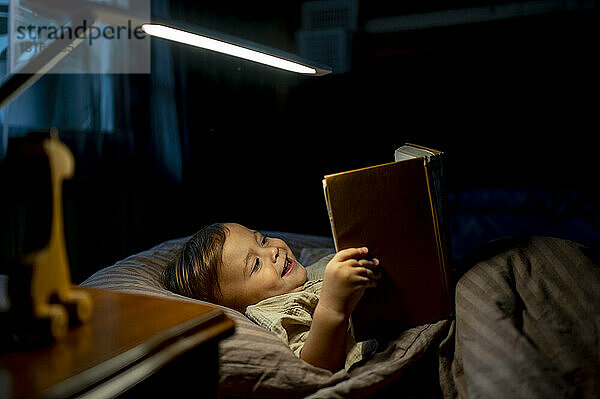 Happy boy reading book lying on bed at home