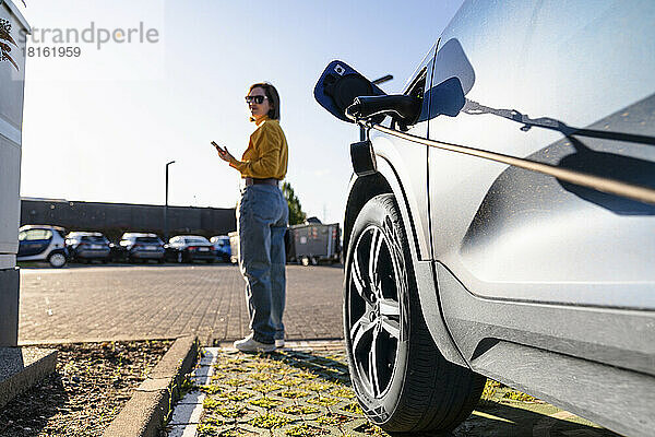 Elektroauto wird an der Station aufgeladen  während eine Frau im Hintergrund an einem sonnigen Tag ihr Smartphone in der Hand hält
