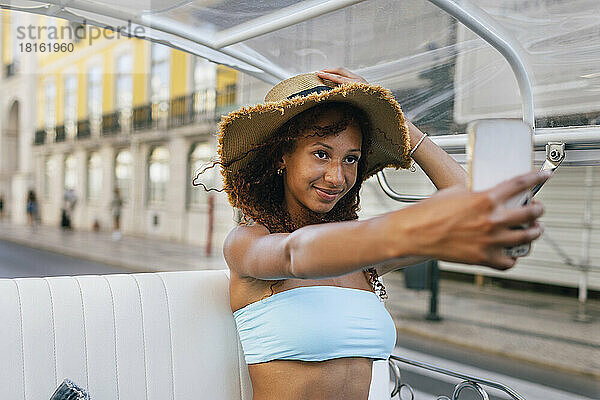 Lächelnde Frau mit Hut macht im Tuk Tuk ein Selfie per Smartphone