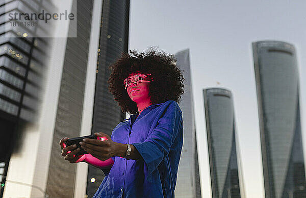 Rotes Licht fällt auf das Gesicht einer Frau  die eine intelligente Brille trägt und ihr Smartphone vor dem Gebäude hält