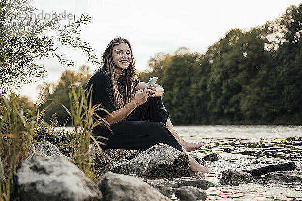 Lächelnde Frau mit Smartphone sitzt am See