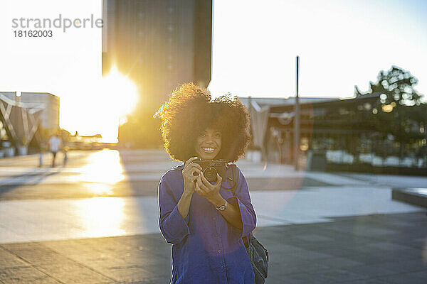 Smiling woman with Afro hairstyle holding camera on footpath