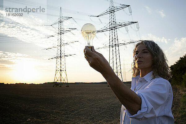 Senior woman looking at light bulb in front of electricity pylons