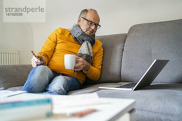 Mann hält Tasse Kaffee und schaut zu Hause auf dem Sofa auf den Laptop