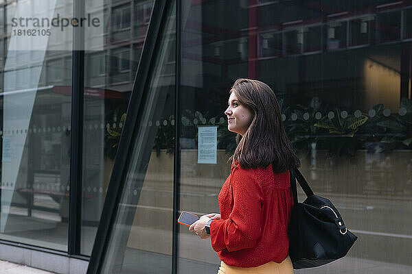 Lächelnde Frau mit Mobiltelefon geht an Glaswand vorbei