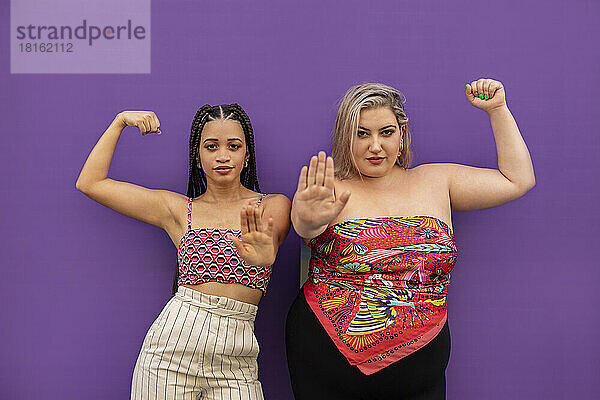 Junge Frauen spielen Muskeln und gestikulieren auf ein Stoppschild vor einer violetten Wand