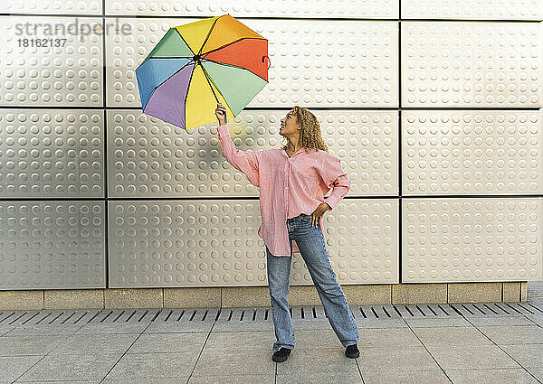 Junge Frau spielt mit buntem Regenschirm vor der Wand