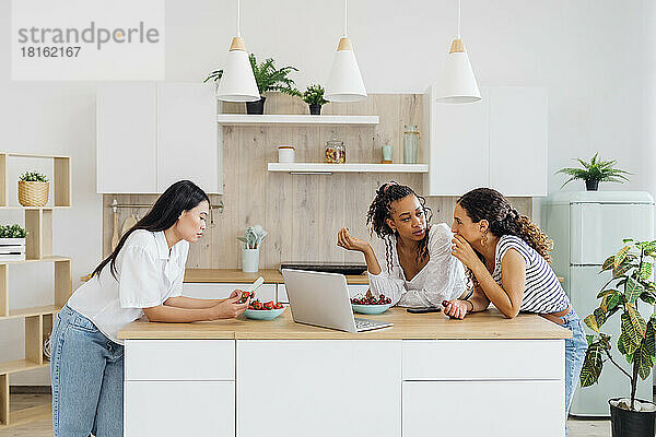 Multiracial friends eating fruits by laptop in kitchen