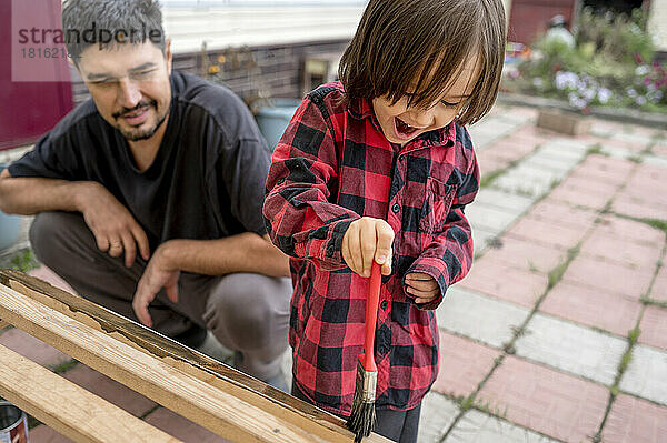 Lächelnder Mann blickt glücklichen Sohn beim Bemalen einer Holzbank an