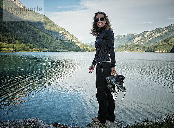 Lächelnde Frau mit Stiefeln steht auf einem Felsen am See