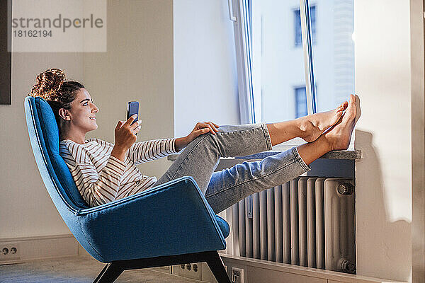Nachdenkliche Frau mit Mobiltelefon entspannt sich zu Hause auf einem Stuhl