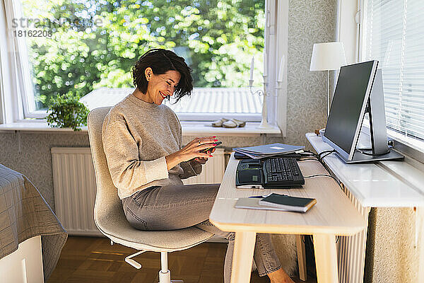 Lächelnde Geschäftsfrau mit Smartphone vor Desktop-PC auf dem heimischen Tisch