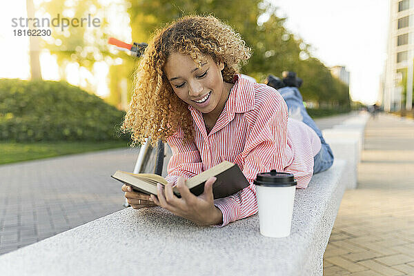 Lächelnde Frau liest Buch auf Bank liegend