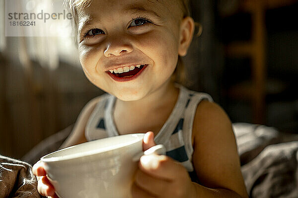 Smiling baby boy with cup in bed at home