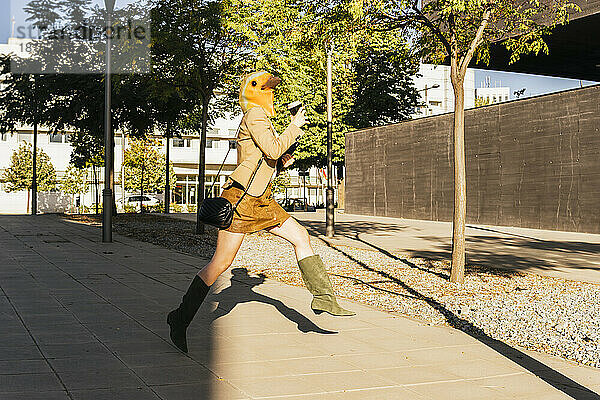Freiberufler mit Vogelmaske springt an sonnigem Tag mit Kaffeetasse auf Fußweg