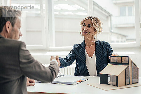 Geschäftsfrau mit Hausmodell schüttelt Kunden im Büro die Hand