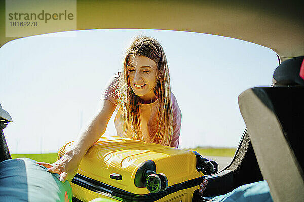 Glückliches Mädchen  das gelben Koffer in den Kofferraum eines Autos lädt