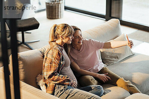 Glückliche Schwester macht Selfie mit schwangerer Frau  die zu Hause auf dem Sofa sitzt