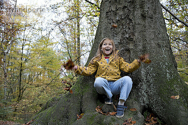 Überraschtes Mädchen  das mit Blättern spielt und auf einem Baumstamm im Wald sitzt