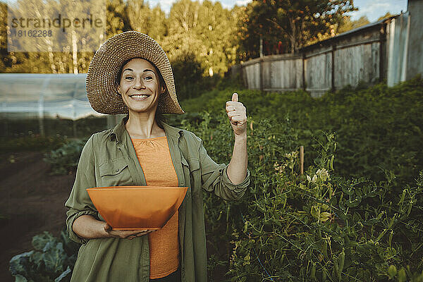 Lächelnde Frau mit Schüssel gestikuliert im Garten mit dem Daumen nach oben