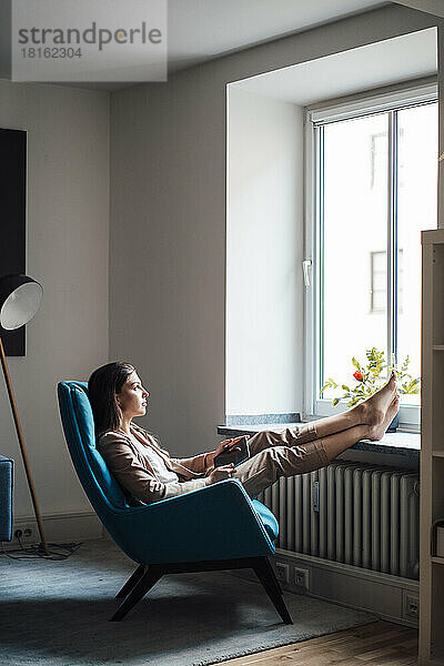Nachdenkliche Geschäftsfrau sitzt zu Hause mit erhobenen Füßen auf einem Stuhl