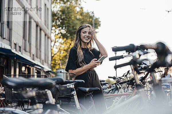 Glückliche junge Frau mit Smartphone steht am Fahrradparkplatz
