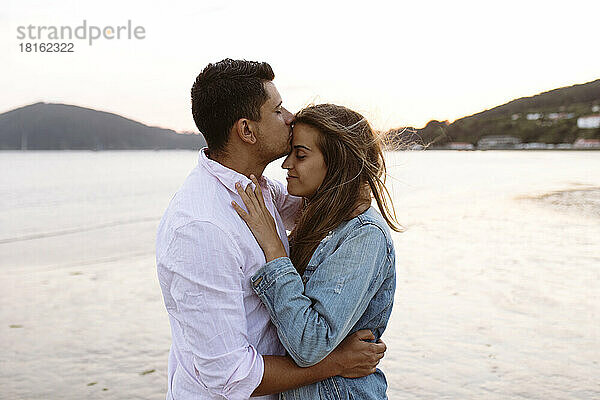 Junger Mann küsst Freundin am Strand auf die Stirn