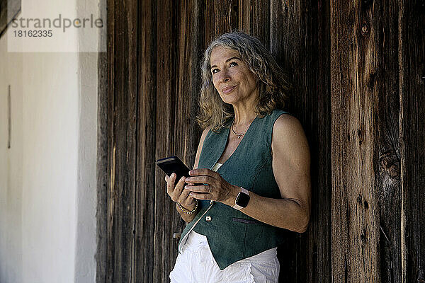 Nachdenkliche ältere Frau mit Mobiltelefon lehnt an altes Scheunentor