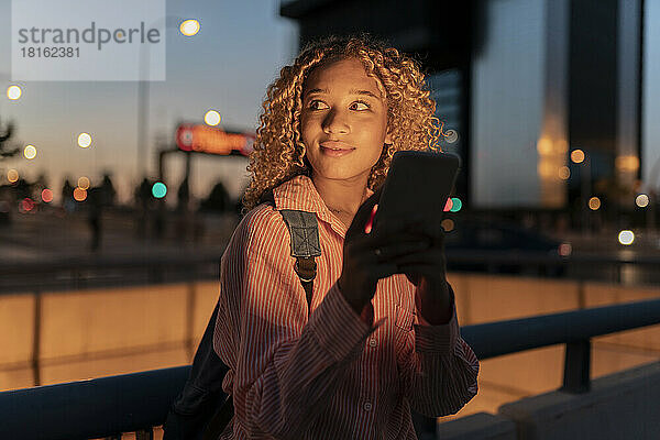Lächelnde junge Frau mit Smartphone am Geländer in der Nacht