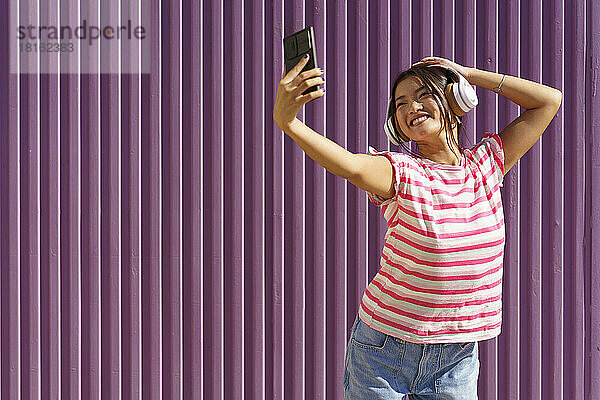 Glückliche junge Frau macht ein Selfie mit ihrem Smartphone vor einer Wellblechwand