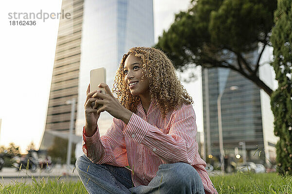 Lächelnde Frau mit Smartphone denkt über Gras in der Stadt nach