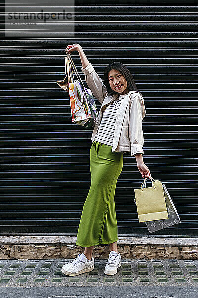 Lächelnde junge Frau mit Einkaufstüten steht vor dem Fensterladen