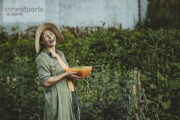 Reife Frau mit grünem Erbsenbehälter lacht über den Garten
