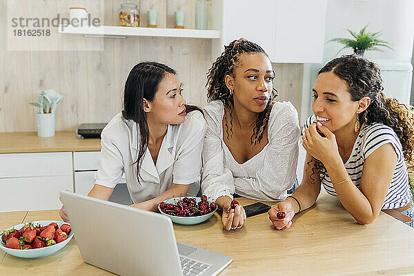 Young women looking at flatmate eating cherry in kitchen