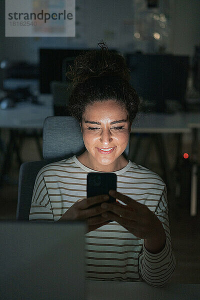 Lächelnde Geschäftsfrau benutzt Mobiltelefon im dunklen Büro