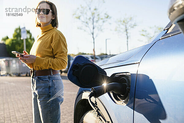 Elektroauto wird an der Station aufgeladen  während eine Frau an einem sonnigen Tag im Hintergrund steht